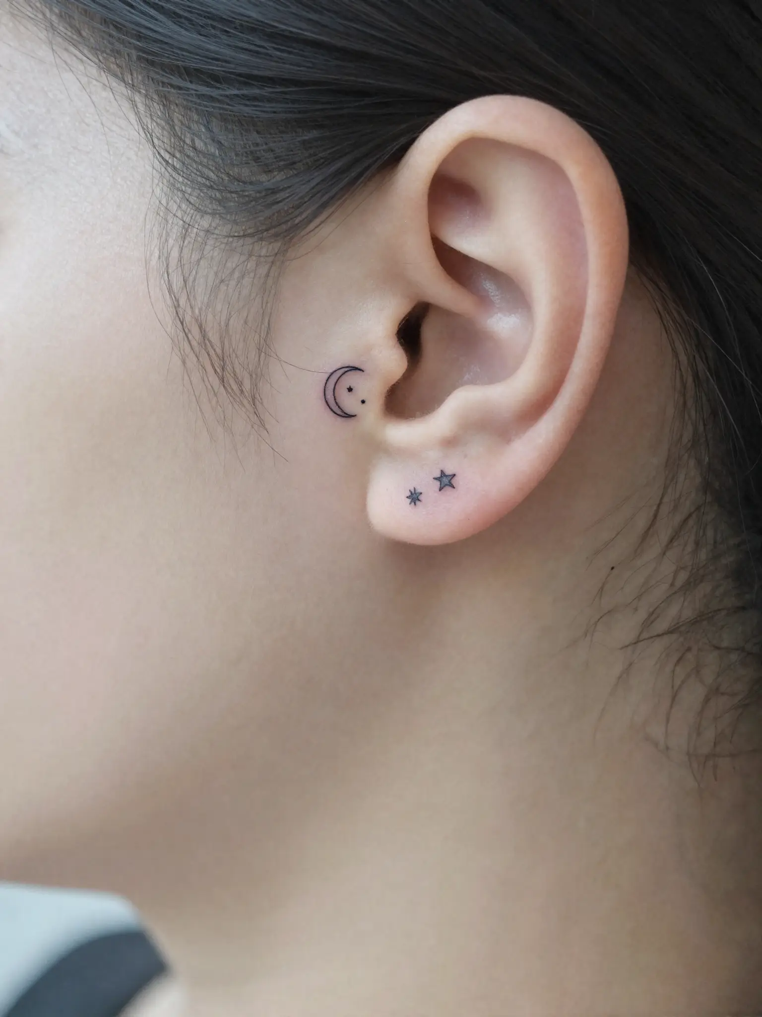 Tatuagem feminina pequena de lua crescente com estrelas atrás da orelha